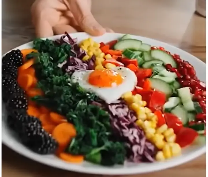 A still from the advertisement: a hand presenting a large white plate of breakfast arranged in strict rainbow stripes: blackberries, carrot, kale, red cabbage, sweetcorn, red pepper, cucumber and pomegranate, with a single fried egg in the middle.